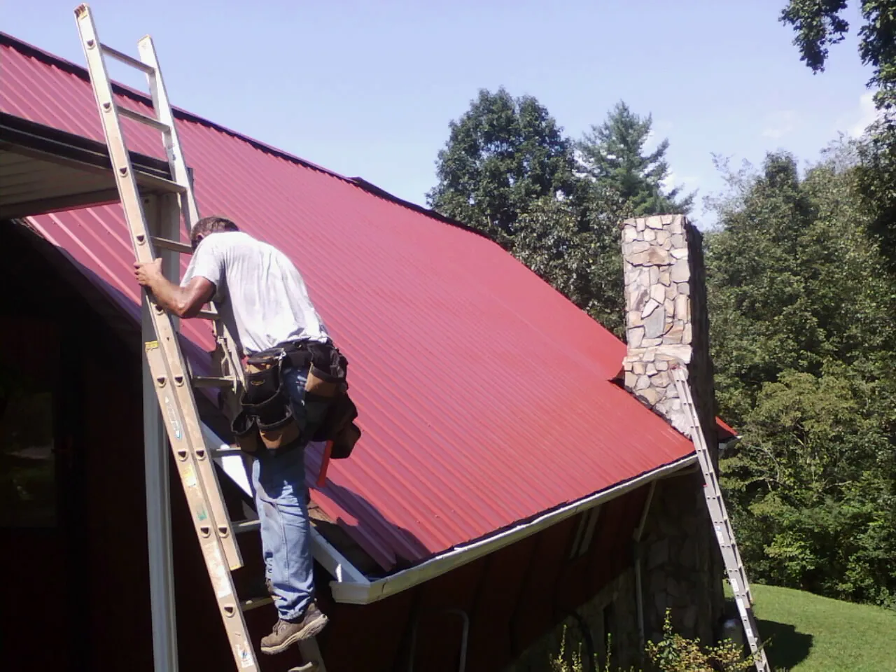 Roofer on a ladder beside a red metal roof and stone chimney — Soffit & Fascia Repair by Roofing Brothers in Alpha